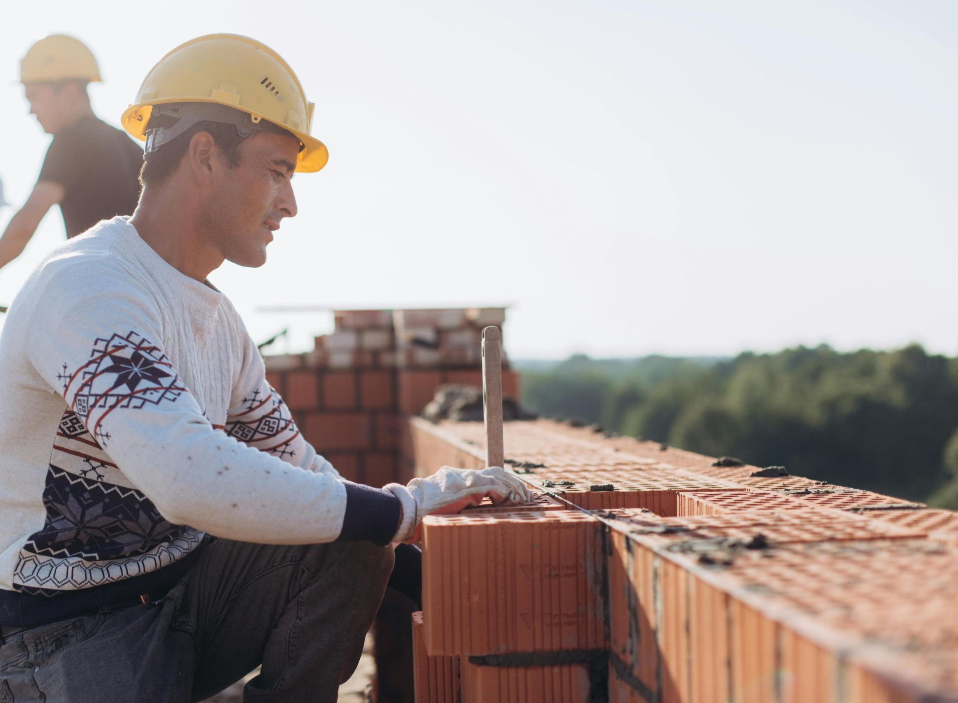 Construction worker laying bricks outdoors in sunny weather with lush greenery in background.